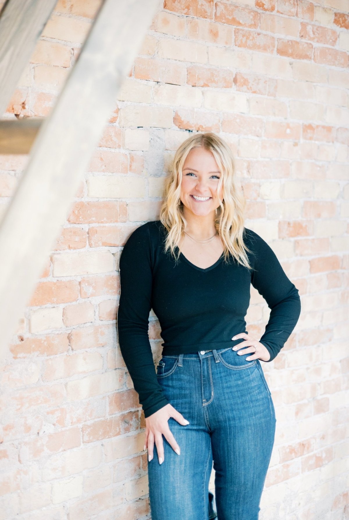 Blonde woman smiling at camera wearing black long-sleeve top and blue jeans, standing against exposed brick wall