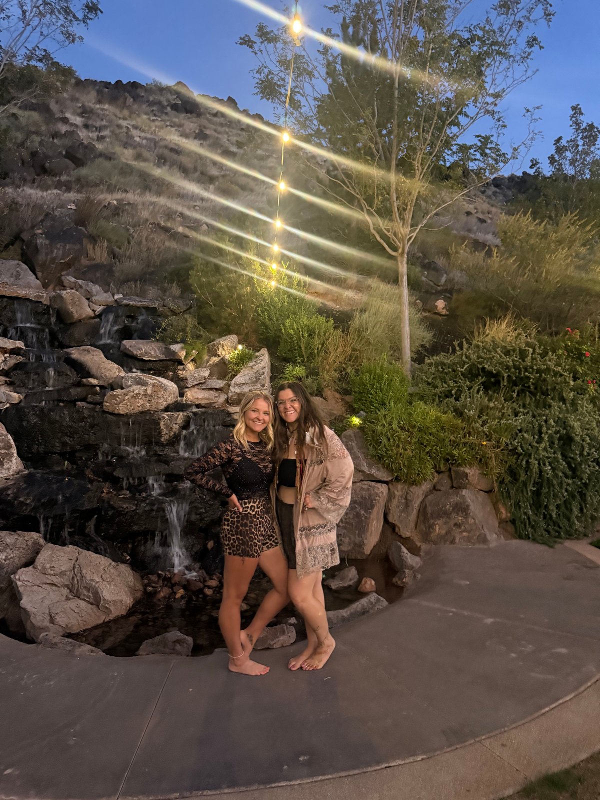 Two women posing together outdoors at night near a small waterfall and illuminated tree in a natural garden setting